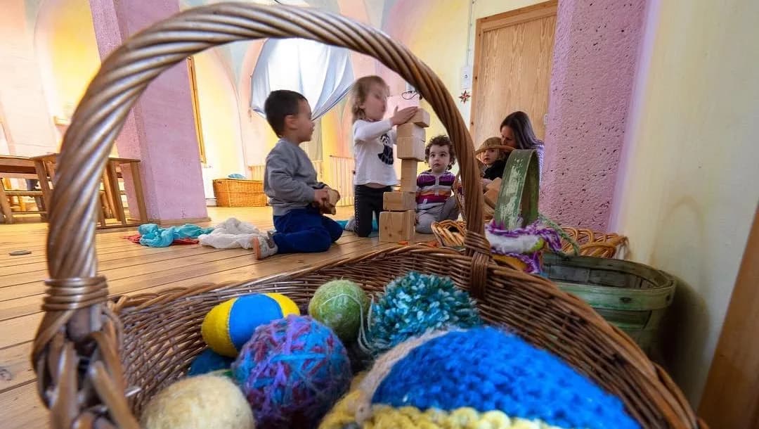 Un cistell de vímet amb pilotes de llana de colors i infants jugant a terra al fons